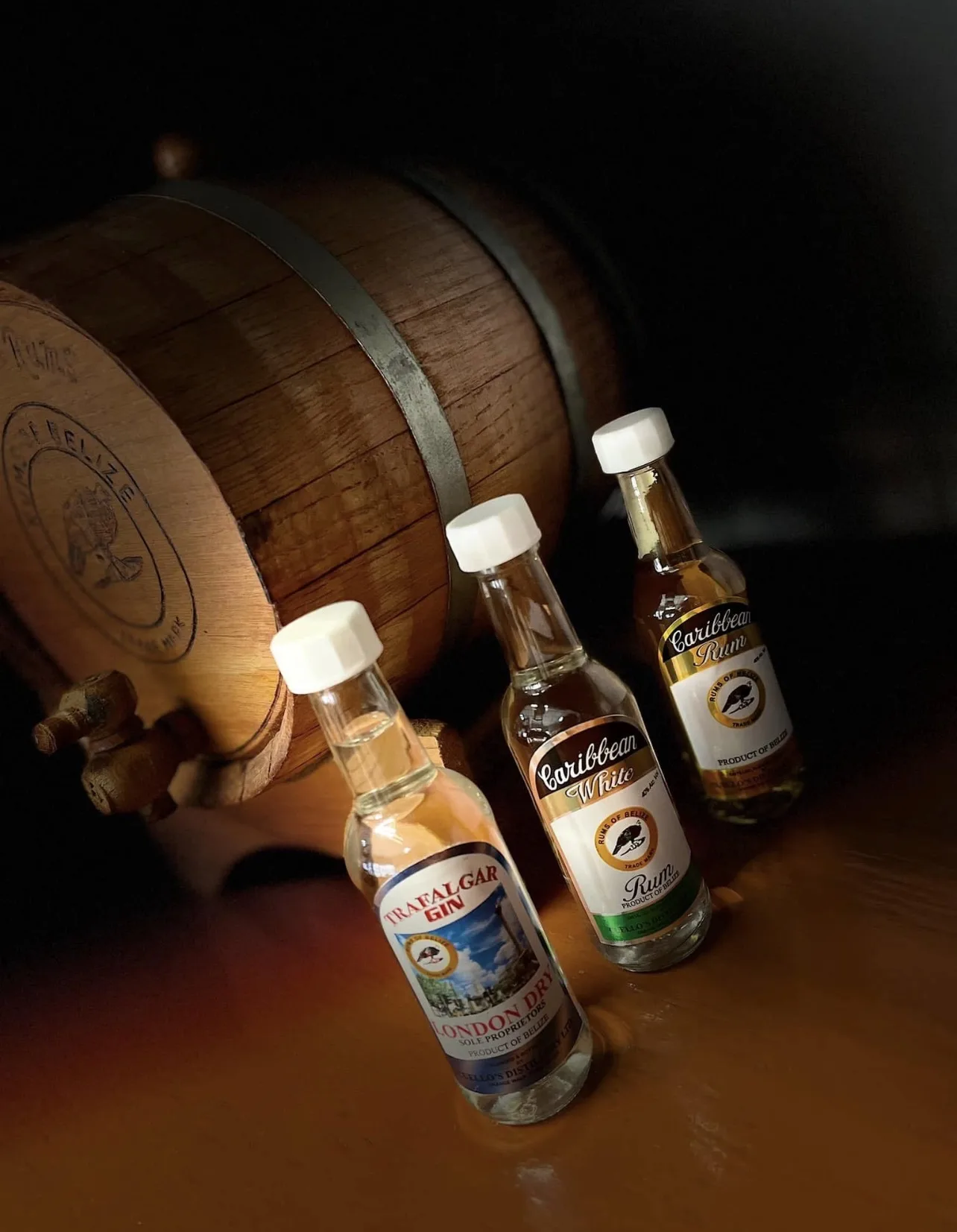 Wooden barrel branded Rums of Belize beside miniature Cuello's bottles in warm light